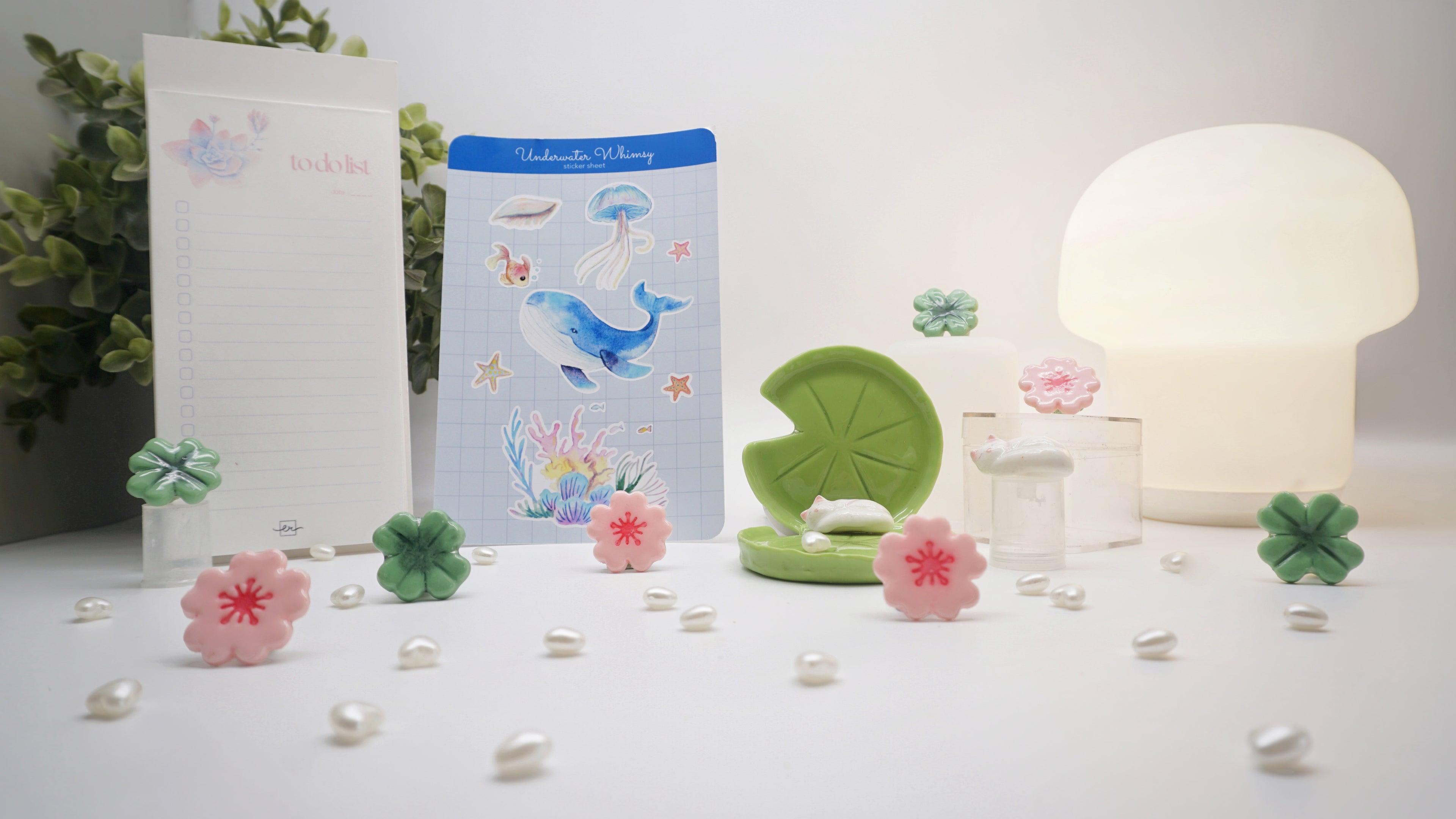 Decorative items including a notebook, stickers, and small plants on a white surface with a lamp in the background.